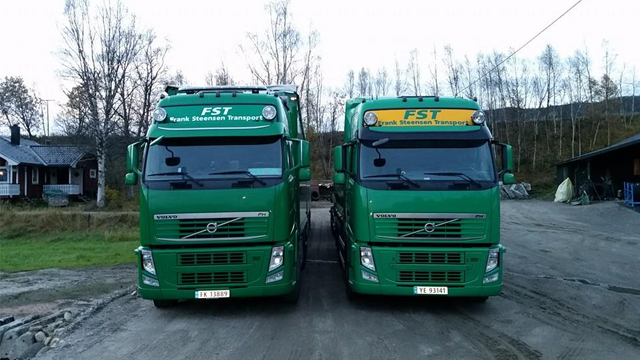 To store grønne lastebiler fra FST Transport parkert på en vei med trær i bakgrunnen.