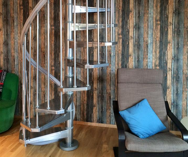 En moderne spiraltrapp i metall ved en vegg med trepanel, med en komfortabel stol med blå pute i nærheten.