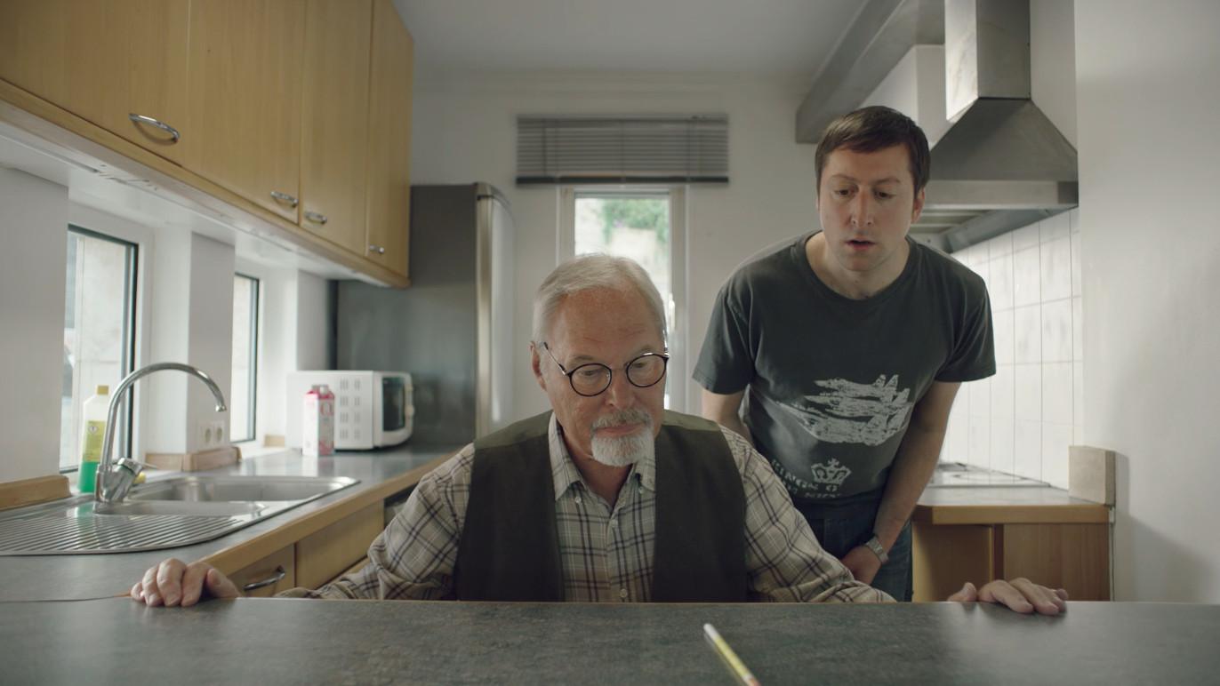 En eldre mann med briller sitter ved kjøkkenbenken, mens en yngre mann står bak ham og ser bekymret ut.