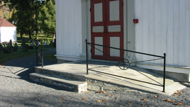 Inngangspartiet til en kirke med en svart jernrekkverk og røde dører, omgitt av grus og gravsteiner.