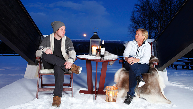 To personer sitter i snøen om kvelden, med en lanterne og vinflaske på et bord mellom dem. Månen lyser opp bak dem.