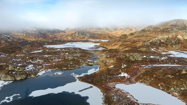 Vakkert fjellandskap med innsjøer, snø og tåke som dekker fjellene i bakgrunnen.