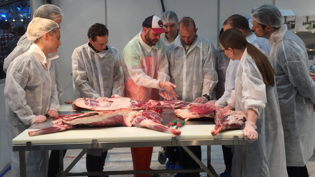 En gruppe mennesker i hvite frakker som lærer om kjøttkutting ved et bord med kjøttstykker.