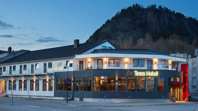 Tinos Hotel med moderne fasade og beliggenhet ved foten av en fjellside, opplyst om kvelden.