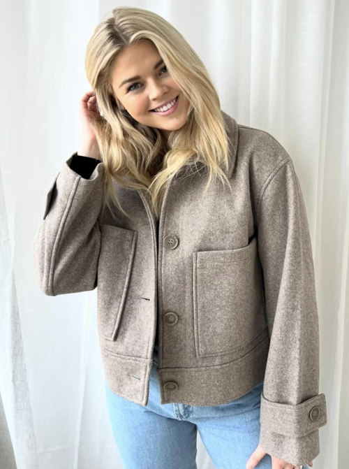 Ung kvinne med langt, lyst hår iført en beige jakke og blå jeans, smilende mot kameraet.
