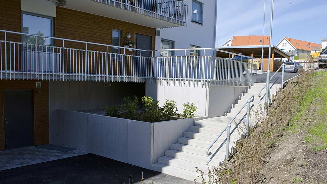 Moderne bygning med trapper og balkong, omgitt av grøntområder og parkeringsplasser.