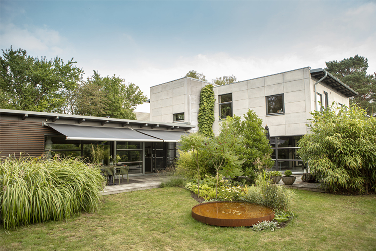 Moderne hus med betongvegg, stor hage med planter og en vannskulptur i midten.