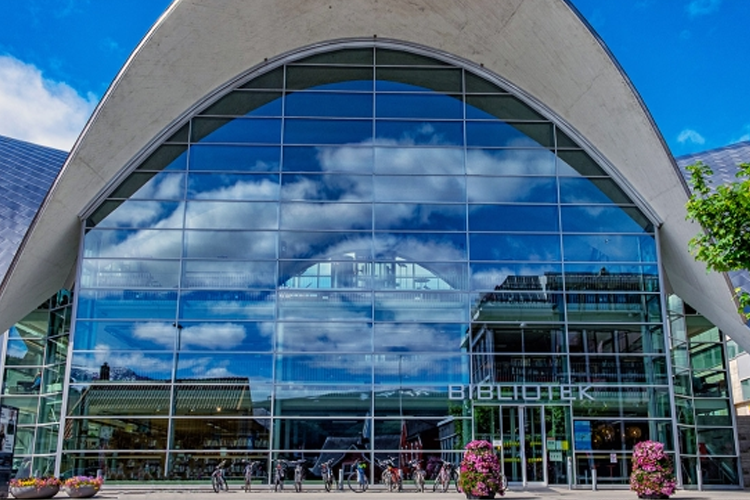 Moderne bibliotek med store glassvinduer som reflekterer himmelen og omgivelsene. Sykkelparkering foran bygningen.