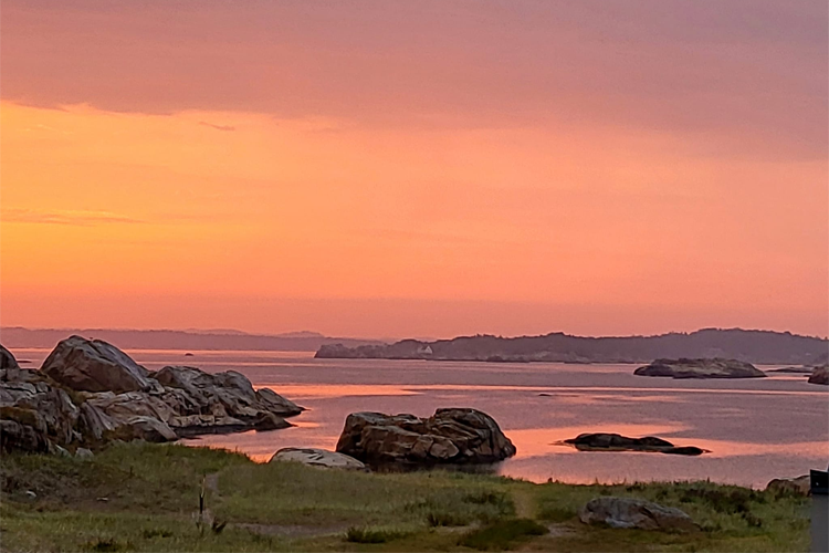 Solnedgang over havet med steiner og øyer i bakgrunnen, fargerik himmel i rosa og oransje nyanser.