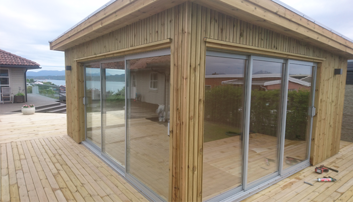 Moderne hytte med store glassdører og trevegg, med utsikt over vannet og terrasse i forgrunnen.
