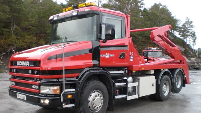 Rød Scania lastebil med kran og tilhenger, parkert på en grusvei omringet av trær.