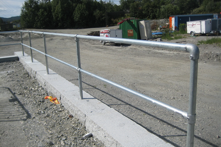 Stålrekkverk på betongfundament ved en byggeplass med grus og containere i bakgrunnen.