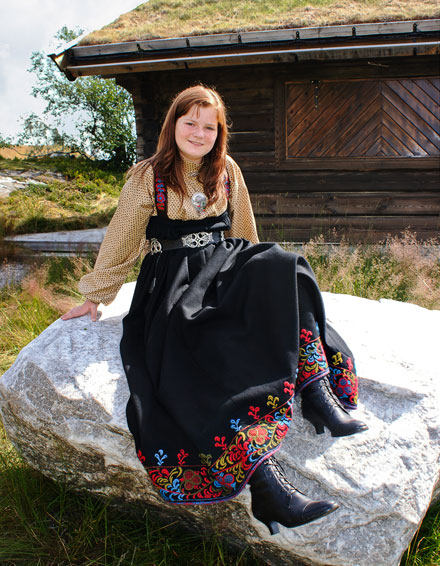 Kvinne i tradisjonell bunad med broderi sitter på en stor stein foran en hytte i naturen.