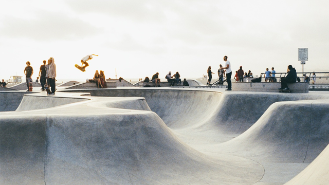 Skater som utfører et hopp i en betong skatepark med folk som sitter og ser på.
