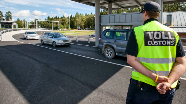En toller i refleksvest overvåker biler som passerer tollstasjonen på en vei omkranset av trær.
