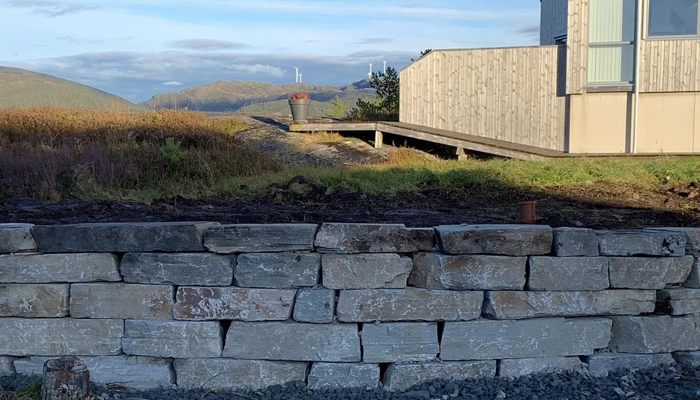 Steinmur med store steiner, med en moderne hytte og fjell i bakgrunnen under en klar himmel.