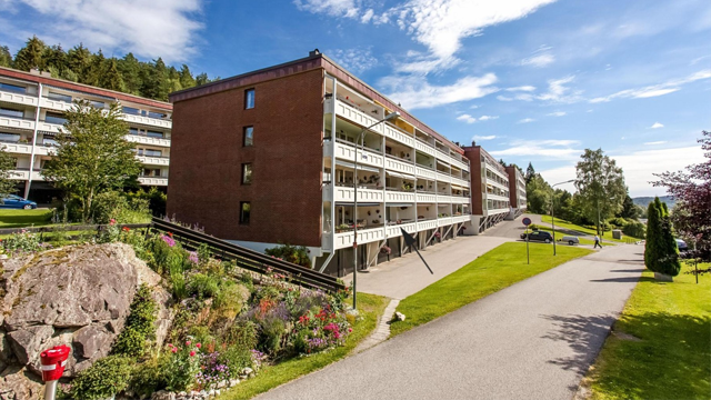 Moderne leilighetskompleks med tre etasjer, omgitt av grøntområde og blomsterbed, under en klar blå himmel.