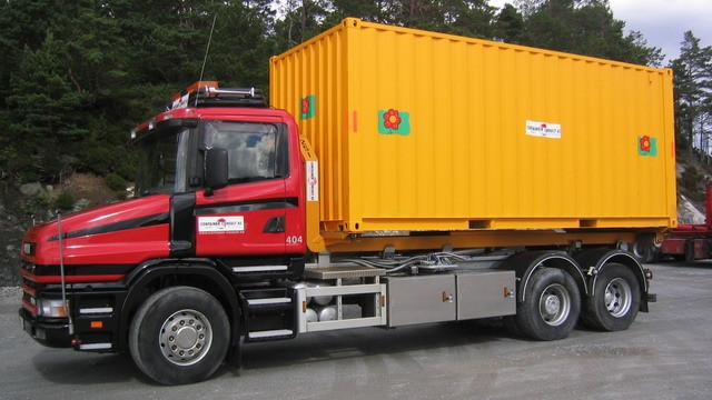 En rød lastebil med en stor gul container på en byggeplass omringet av trær.