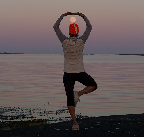 En person i yoga-posisjon med månen over hodet, ved havet under solnedgang.