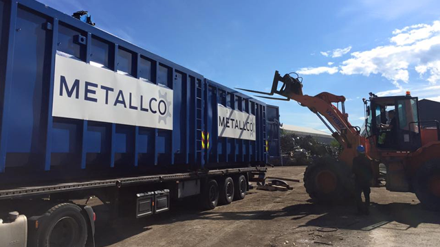 En gravemaskin laster metallcontainere på en lastebil med Metallco-logo. Solfylt dag på en byggeplass.