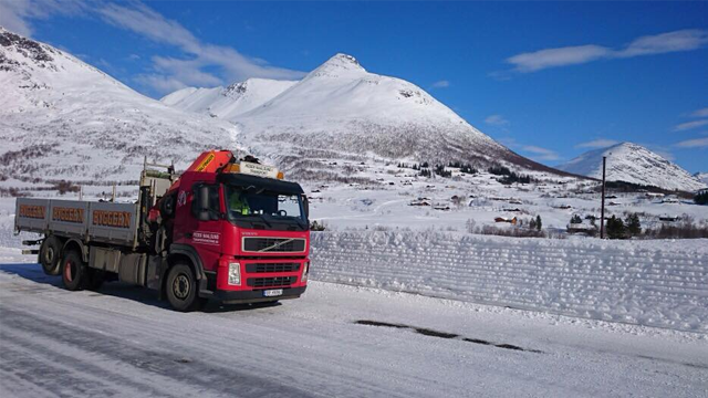 En rød lastebil står på en snødekt vei med fjell i bakgrunnen under en klar blå himmel.