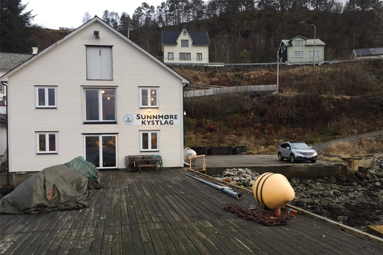 Hus ved vannet med skilt for Sunnmøre Kystlag, båter og en bil parkert i nærheten.