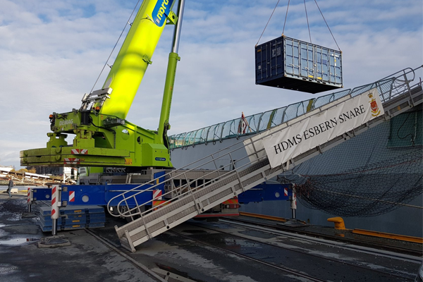 En stor kran løfter en container over en båt ved havnen, med en rampe for lasting og lossing.