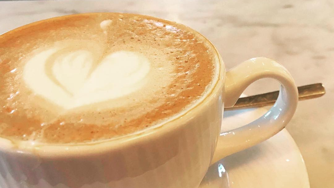 Kopp med latteart i form av et hjerte, plassert på en hvit tallerken med en skje ved siden av.