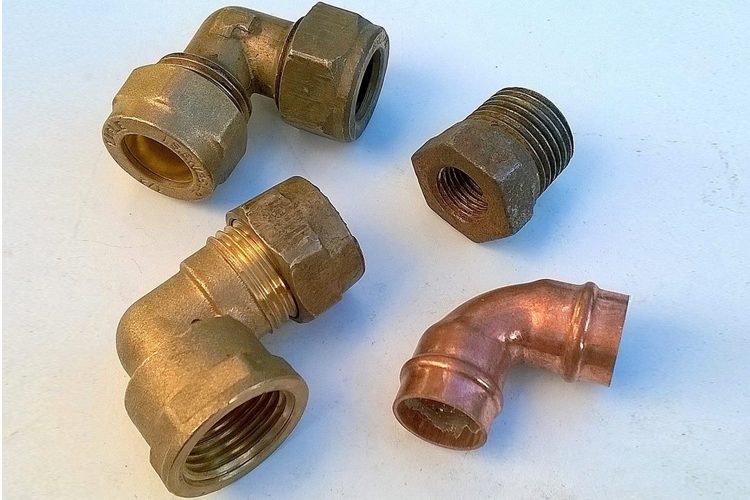 Fire rørfittings i messing og kobber, inkludert vinkelfittings og muttere, på en lys bakgrunn.