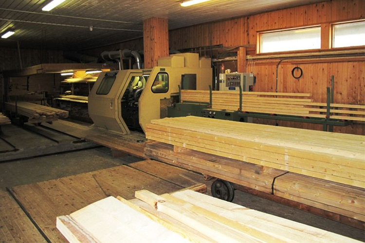 En trebearbeidingsmaskin i en verksted med lange planker stablet på en vogn. Veggene er av trepanel.