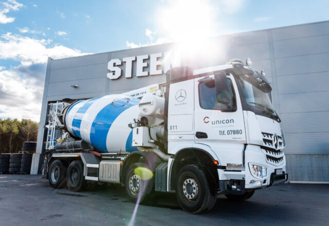 En betongbil fra Unicon med blå og hvite striper står foran en bygning med teksten "STEEL" på fasaden.