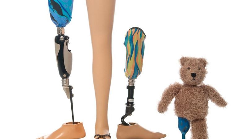 En dukke med proteser og sko står ved siden av en kosebamse med blå fot.