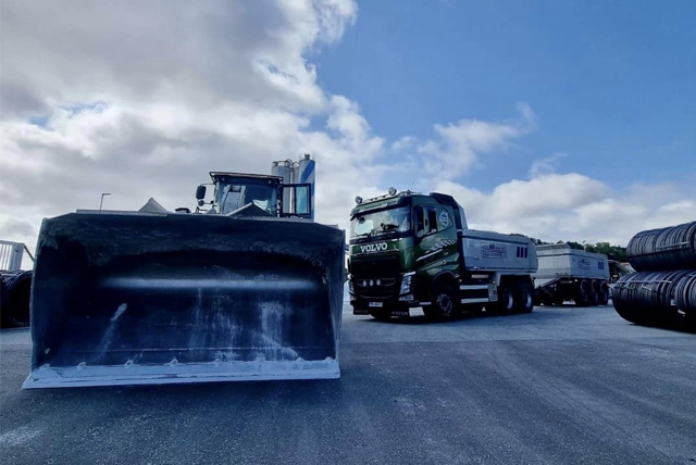 To lastebiler med snøploger og tilhengere på en parkeringsplass under en blå himmel.
