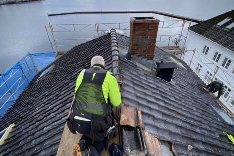 Taklegger som arbeider med takstein på et hus med utsikt over vannet. Byggeplass med stillas i bakgrunnen.