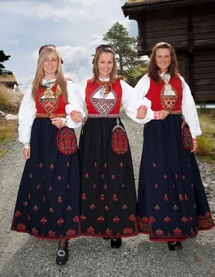 Tre kvinner i tradisjonelle bunader med røde og svarte farger, står sammen på en grusvei med bygninger i bakgrunnen.