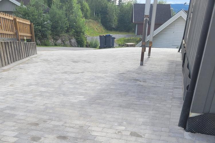 En grå steinlagt innkjørsel med trær og hus i bakgrunnen, samt søppeldunker til venstre.