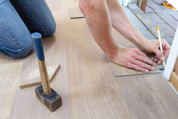 En person som legger gulvplanker, bruker en hammer og måler med blyant på et tregulv.