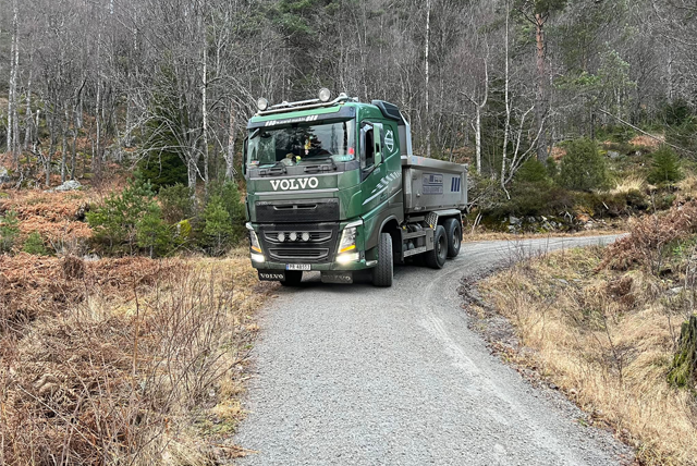 En grønn Volvo lastebil kjører på en svingete grusvei omringet av trær og busker.