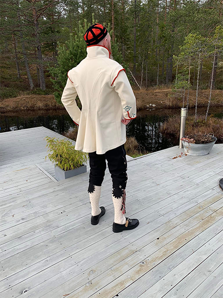 En person med tradisjonell norsk drakt står på en tredekk med skog og vann i bakgrunnen.