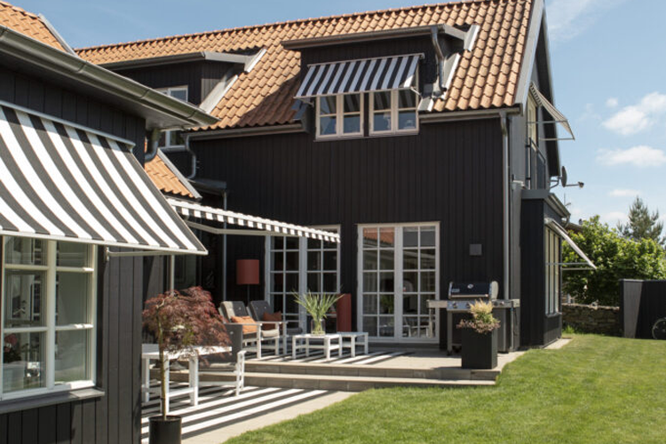 Moderne svart hus med rød takstein, terrasse med møbler og markiser, omgitt av grønt gress og planter.