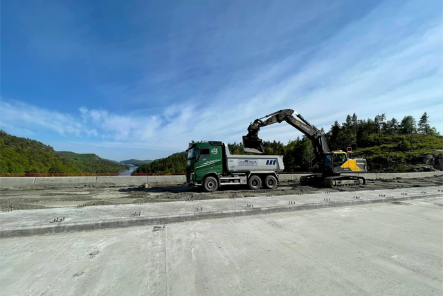 Grønn lastebil og gravemaskin arbeider på en byggeplass med fjell og elv i bakgrunnen.
