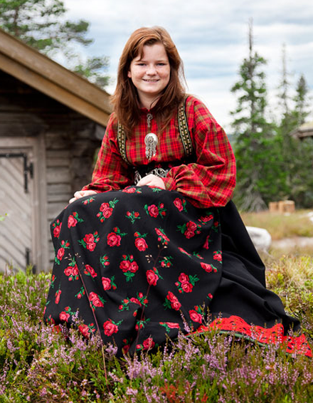 Ung kvinne i tradisjonell bunad med rød bluse og svart skjørt med rosemønster, sitter på blomstereng.