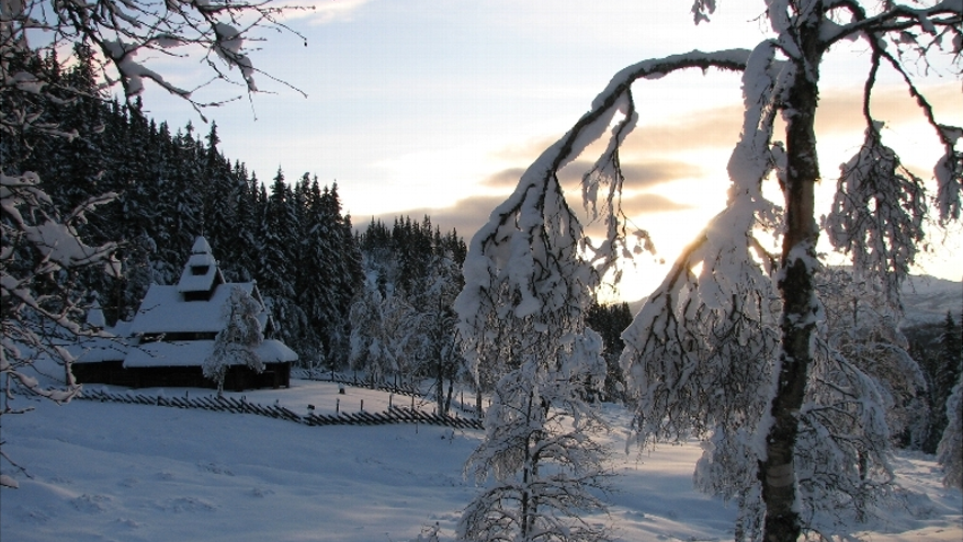 Vinterlandskap med snødekt trehus, omringet av snødekte trær og en solnedgang i bakgrunnen.