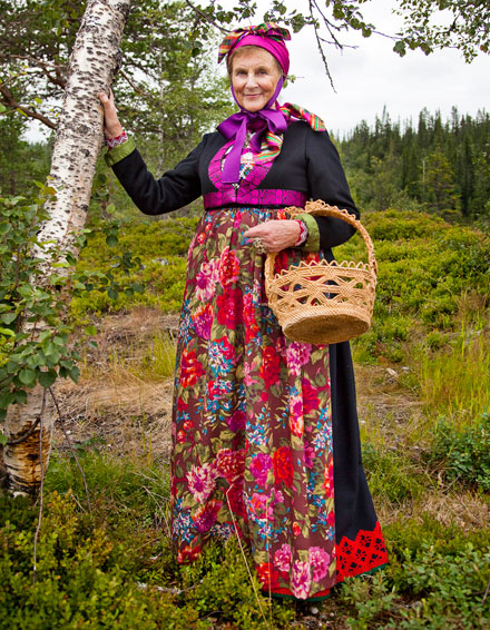 Kvinne i tradisjonell bunad med blomsterdesign, holder en kurv, står ved et bjørketre i en grønn eng.