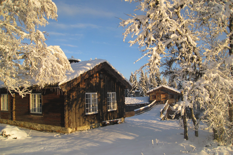En koselig hytte dekket av snø, omgitt av snødekte trær og en klar blå himmel.