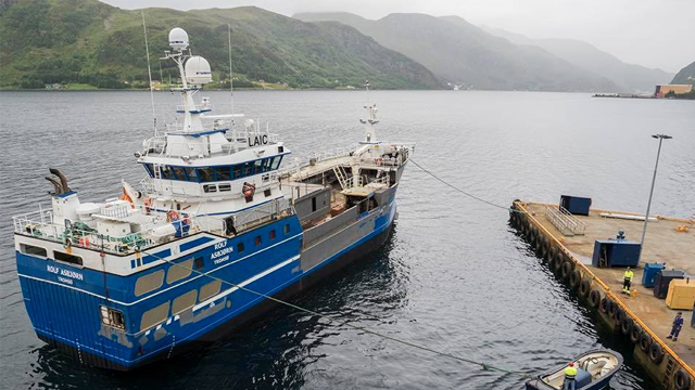 Fiskebåt i havn med fjell i bakgrunnen, fortøyd ved kaien med arbeidere i nærheten.