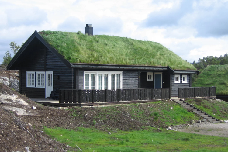 En svart hytte med gressdekket tak, plassert i et naturskjønt område med grønt landskap.