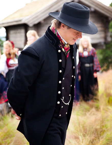 Ung mann i tradisjonell norsk bunad med hatt, går med hendene bak ryggen, omgitt av folk i festklær.