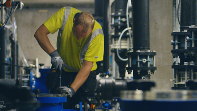 En mann i gul arbeidsdrakt jobber med verktøy i en industriell setting med rør og pumper.