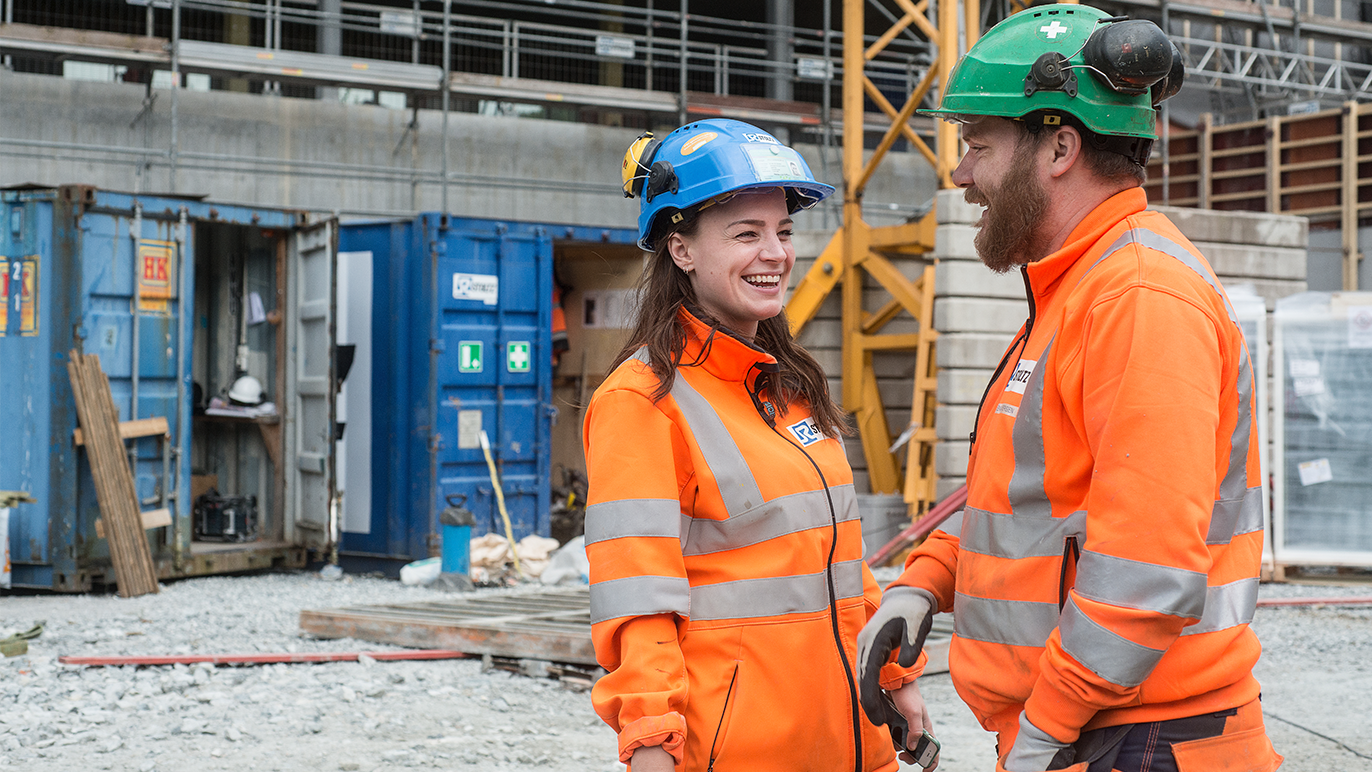 To personer i arbeidsklær og hjelmer smiler og snakker på en byggeplass med containere i bakgrunnen.